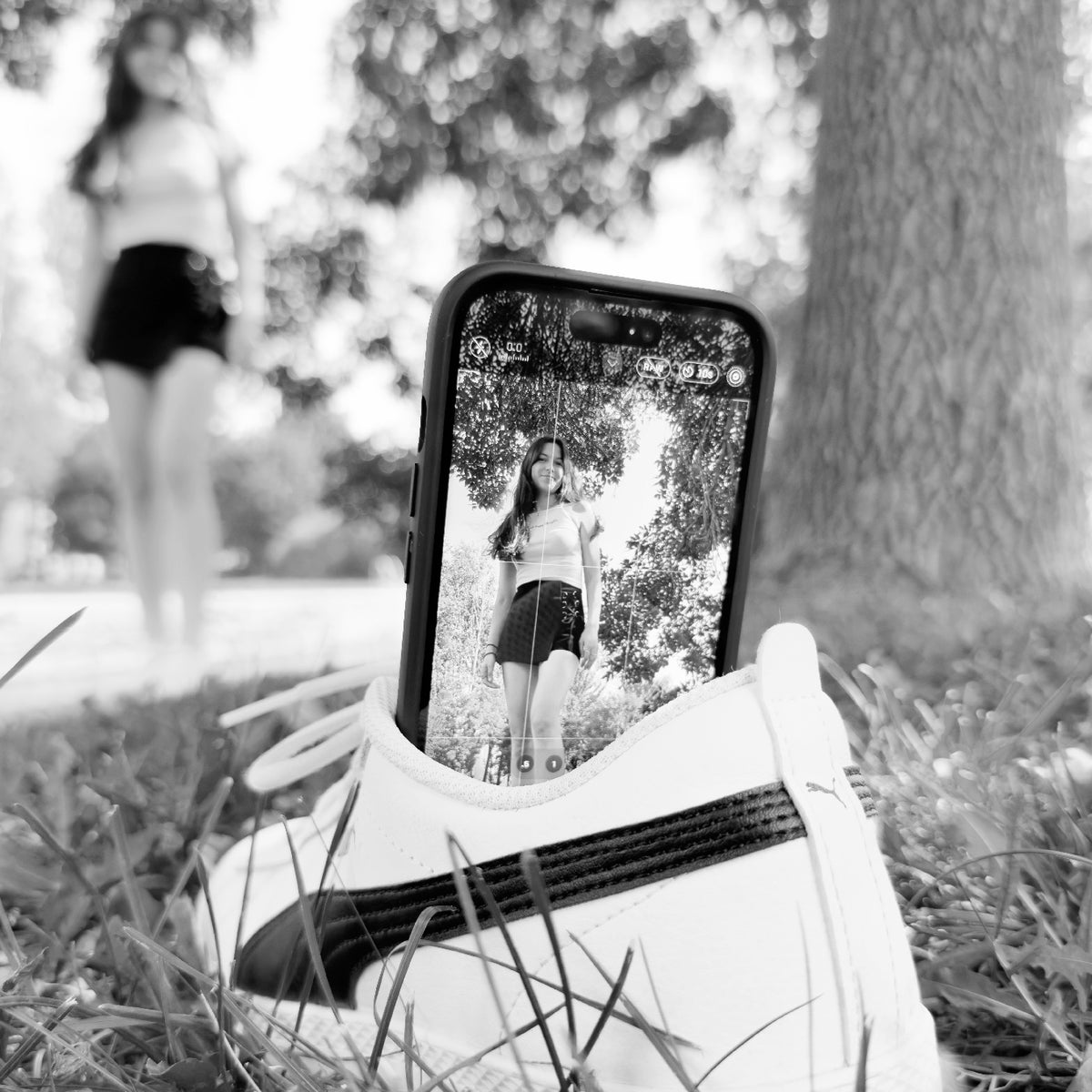 Black-and-white photo: a phone propped against a sneaker in the grass, filming a woman in a park