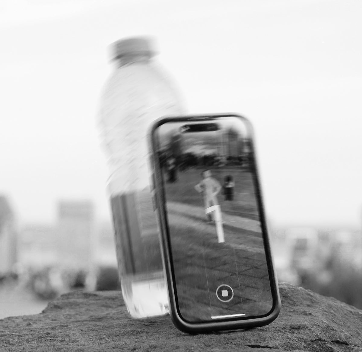 Black-and-white photo: a phone leaning on a water bottle on a rock to take a picture