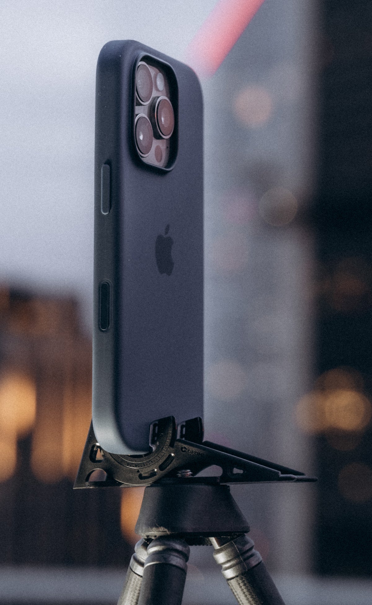 A phone in a dark case on the Pocket Tripod atop a camera tripod, city lights behind