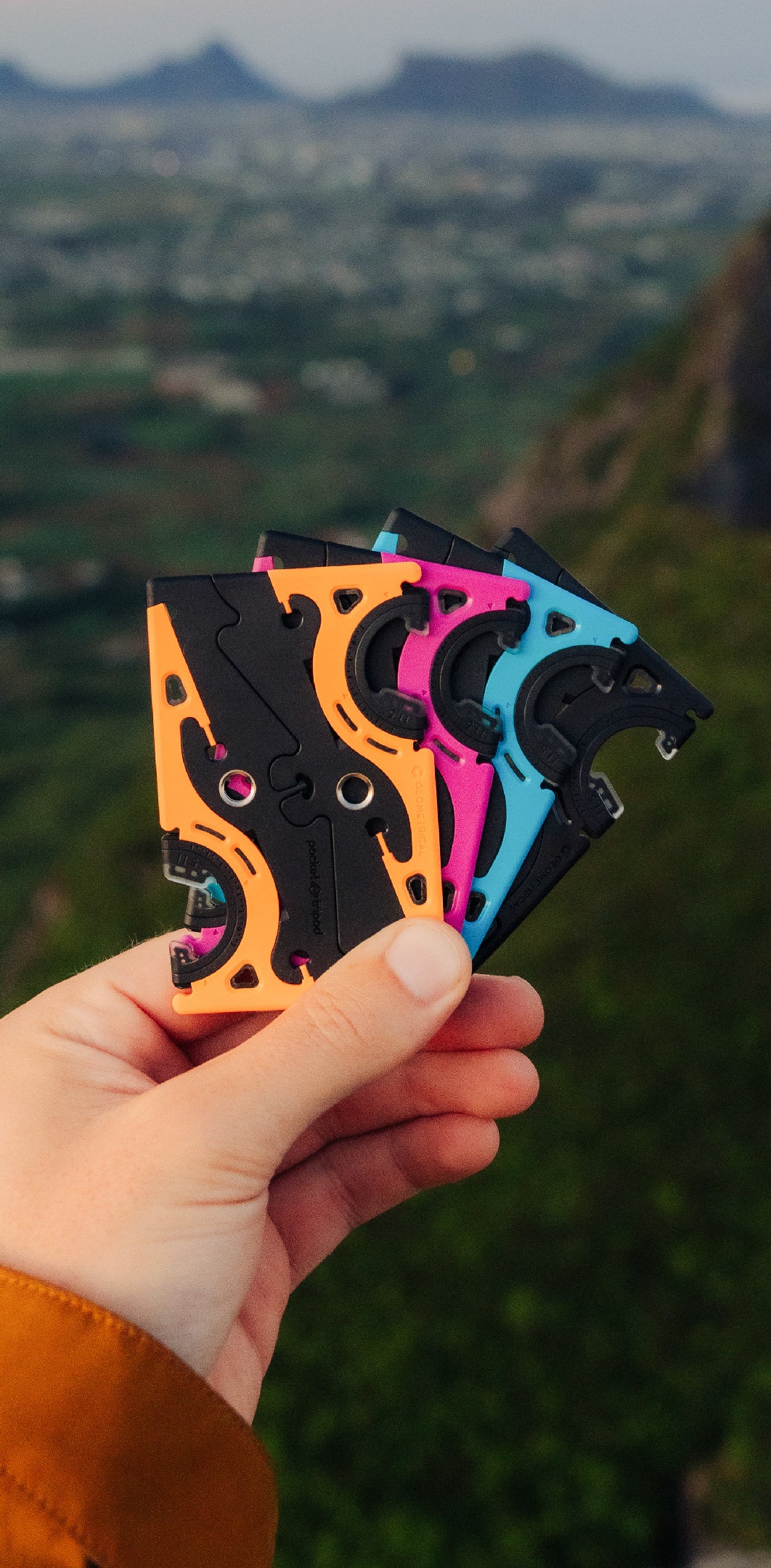 A hand fanning out Pocket Tripods in orange, pink, blue and black above a green valley