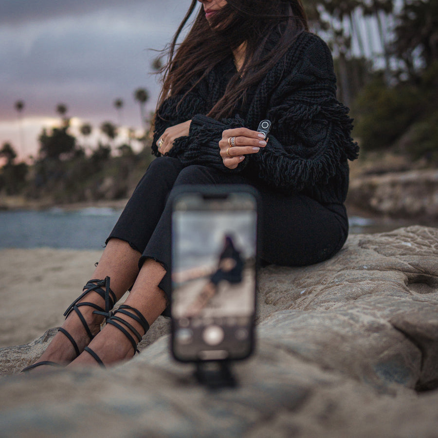 A woman sitting on rocks at dusk holding the shutter remote, with her phone on a Pocket Tripod in the foreground framing her
